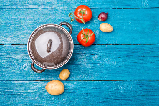Photo of iron pot, tomato, potato, onion on blue wooden background.