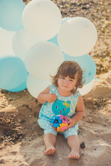 Baby cute girl with blond hair and pink apple cheek enjoying summer time holiday posing on sand beach sea side with blue white balloons wearing casual kids clothes