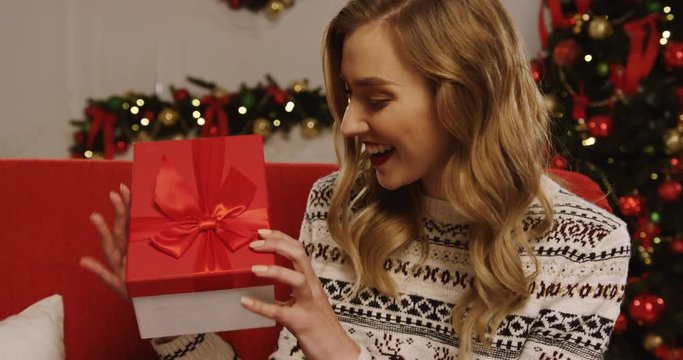 Attractive blonde young woman shaking her Christmas present and opening it with surprised eyes while sitting on the red sofa in the room on the Christmas tree background. Indoor. Close up. Portrait
