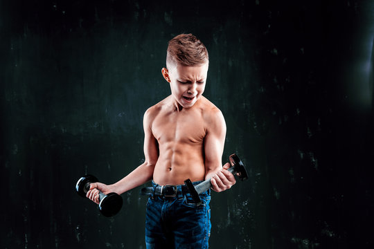 Bare-chested boy in jeans lifts heavy dumbbell while standing on dark wall background.