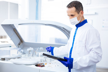 Cutting edge equipment. Attractive focused  male scientist reading instruction for innovative centrifuge machine  while holding tablet and standing in lab
