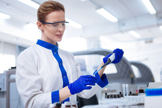 Caution with chemicals. Blond attractive female scientist   looking down and  holding pipette when posing against blurred background