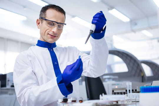 Great sample. Waist up of tranquil enthusiastic male lab assistant  who taking sample from the glassware with blue chemical placed there when standing on the blurred background 