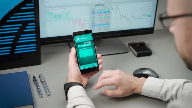 The guy in the office uses his bitcoin purse on the smartphone. In the frame of the hands on the phone, the man enters his electronic account and looks through the transactions.