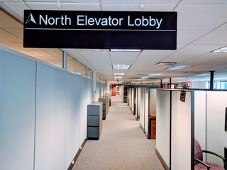 Empty office cubicles along a walkway with an overhead sign indicating the location of elevators.