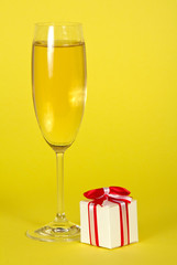 A glass of sparkling wine and small souvenir decorated with ribbon on yellow background