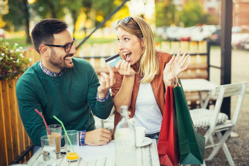 Woman sitting in cafe with handsome man and colorful shopping bags, cup of coffee, holding credit card. On line shopping concept. Beautiful couple smiling.