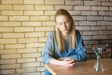 Sad blonde girl in blue blouse sitting at the table in a cafe, listening music with headphones