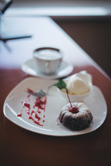 Focus close picture of delightful sweet fancy looking desert chocolate muffin with cherry and cup of coffe on white plate on wooden table in contemporary restaurant caffe