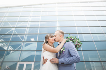 Lovely happy wedding couple, bride with long white dress posing near modern building