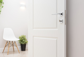 The open white interior doors. Modern chrome handle and lock with key. Wall lamp, chair, and green plant in the background