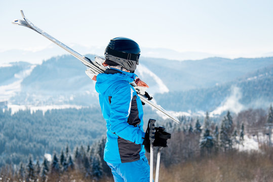 Rear view female skier holding skis on the shoulder, wearing blue ski suit and black helmet, resting on top of the mountain observing nature at ski resort on a beautiful sunny winter day