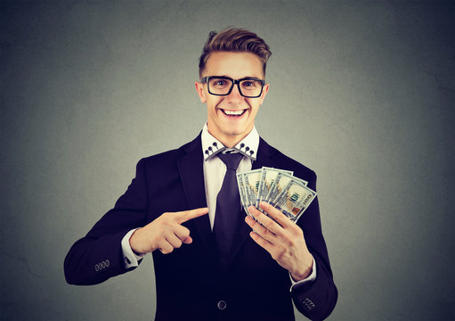 Successful young business man holding money dollar bills in hand isolated on gray background. Positive emotion facial expression. Financial reward concept 