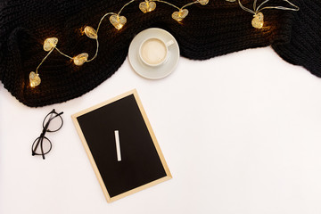 A cup of coffee, a small black wooden board and a white chalk with new years goals. Scarf and Christmas lights on white background.
