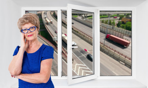 Happy smiling senior woman stands inside near threefold pvc window pane with hoisy highway with cars on background