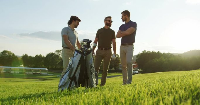 Three handsome male golf players talking and holding the golf bag on the nice sunny day on the golf field. Outside. Shot from below
