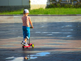 A little girl in colorful summer clothes is riding a children's scooter among puddles on a tile floor.