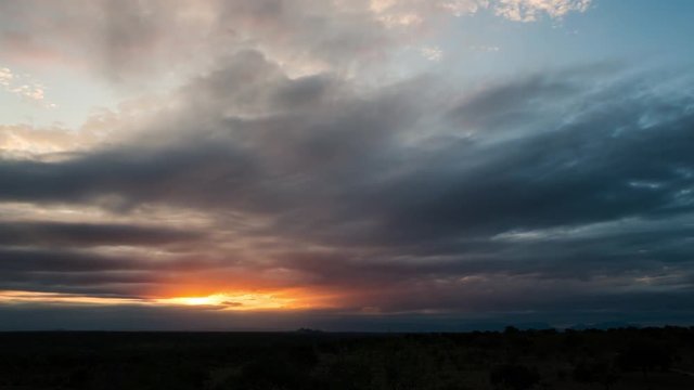 A tilt down timelapse at sunset with dramatic and dark clouds as the orange sky fades to dark blue over a wide bushveld landscape. 