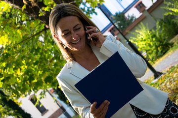Happy businesswoman talking on cell phone while reading reports in nature.