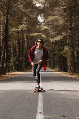 Obraz premium A young hipster in a cap and plaid shirt is riding his longboard on a country road in the forest