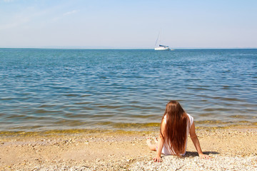 Fototapeta na wymiar A girl with long hair in white clothes is sitting on the rocks on the bank in front of a river and a white yacht on a sunny summer day.