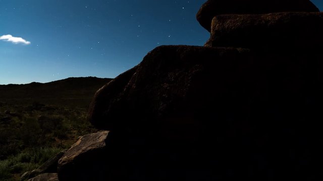 A vertical linear timelapse (moving up) of a rocky moonlit landscape with abstract shadows and rocks, scattered clouds and stars against a blue night sky with a slow focus pull from the foreground to the distant landscape. 