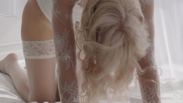 Sexy woman in white underwear flaps long blond hair on bed. Passionate blonde with a bright make-up seductively moves on her knees.