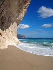 Ein blaues Meer im Hintergrund die Berge, vorne ein unberührter Sandstrand.  Auf der linken Seite eine überhängende Klippe aus Sandstein