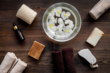 Bath in bowl with tropical flowers for foot spa, pumice stone, soap and oil on dark wooden background top view