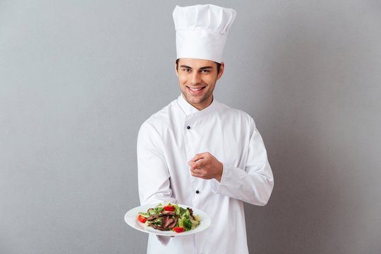 Cheerful young cook in uniform holding salad pointing to you.