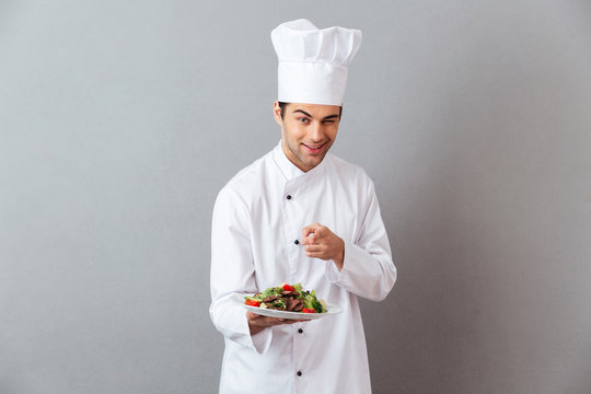 Cheerful young cook in uniform holding salad pointing to you.