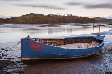 An dilapidated boat resting on the mud flats of the River Urr in Kippford waiting for the tide to return to refloat again. The autumn colours can be seen in the trees on hills on the opposite bank