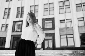 Portrait of a fabulous young successful woman in white blouse and broad black pants posing on the stairs with a huge white building on the background. Black and white photo.