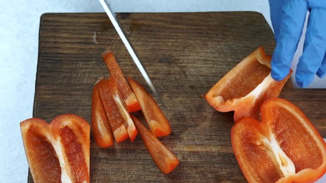 The quartets of red pepper is being sliced for smaller pieces by chief. The kitchener wears blue glove on the hand and cutting paprika with big steel knife on wooden board to cook the salad in the
