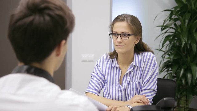 In office woman in glasses holds an interview with man about hiring. Adult headmaster is interviewing young guy for vacant job in a large company.