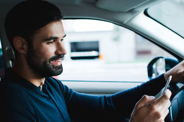 Going for a date. Side view of handsome young man using his smart phone with smile while sitting in car