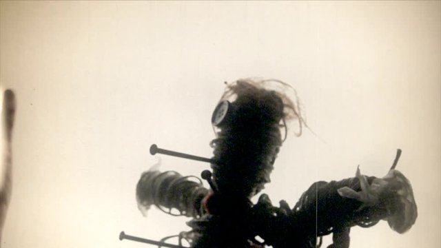 Fake 8mm amateur film: the scay silhouette of a cursed voodoo doll, pierced by big rusty nails, over a white background.
