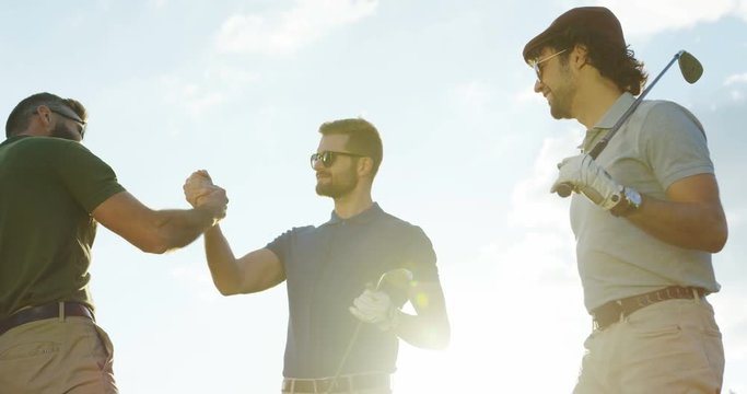 Portrait shot of male friends with golf clubs meeting outside and shaking hands. Sunny sky background