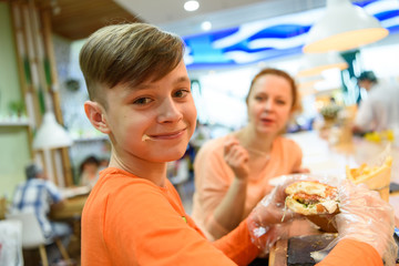 In cafes stained cheese happy son eats with gloves on a huge sandwich. Mom tells how to eat.