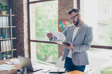 Focused concentrated accountant, fashionable brunet bearded guy is looking at the report, stands in...