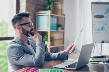 Creative occupation, making decision, looking at the pantone. Side view profile shot of stylish bearded male designer with mustache in trendy specs and grey formal jacket, chooses colors for project