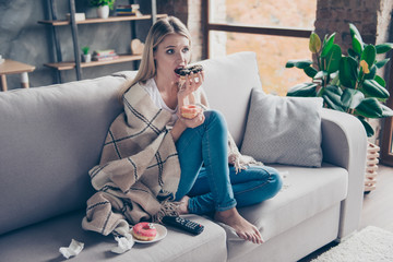 Beautiful blonde woman sitting on couch in living room under blanket eating chokolate donat...