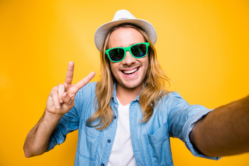 Close up portrait of happy hipster man in summer glasses with beaming smile making selfie and showing v-sign in camera standing over yellow background