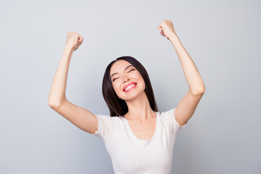 Close up of overjoyed, successful young manager closed her eyes  with two hands raised and celebrating victory while standing in front of grey background