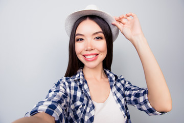 Selfie mania. Close up of confident brunette student  with beaming smile  taking a self-portrait  on her mobile phone and touching her cap over grey background