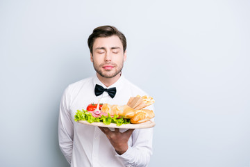 Concept of always fresh tasty and qualitative food in a restaurant industry. Happy smiling waiter with closed eyes is enjoying the aromatic smell of the dish on a tray, isolated on grey background