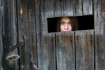 Young woman locked in a wooden barn, looking out of the window slit. Violence against women concept.
