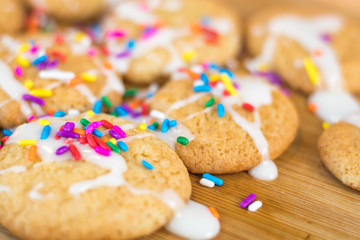 Freshly baked sugar cookies with white icing and rainbow colored sprinkles on wooden board, selective focus