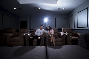Interracial couple on a movie date in a living room with a home theater system with seats. They are drinking red wine on glass. They are watching a movie or television.