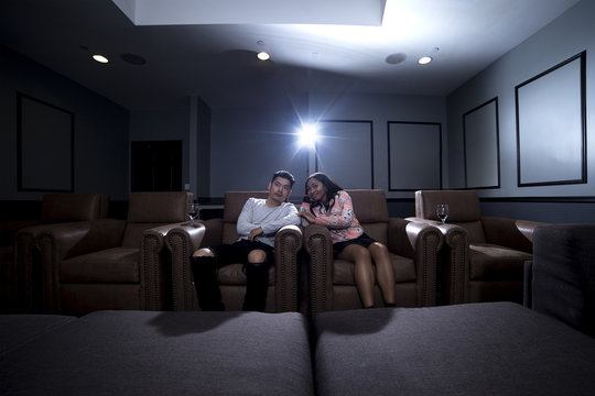 Interracial Couple On A Movie Date In A Living Room With A Home Theater System With Seats.  They Are Drinking Red Wine On Glass.  They Are Watching A Movie Or Television.